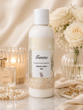 A bottle of Femme Cloud Lotion 4oz centered on a marble surface, surrounded by soft cream satin fabric, white roses in a glass vase, pearl strands, a lit candle, and gold jewelry. The product bottle is white with a beige-and-white label and a white cap.